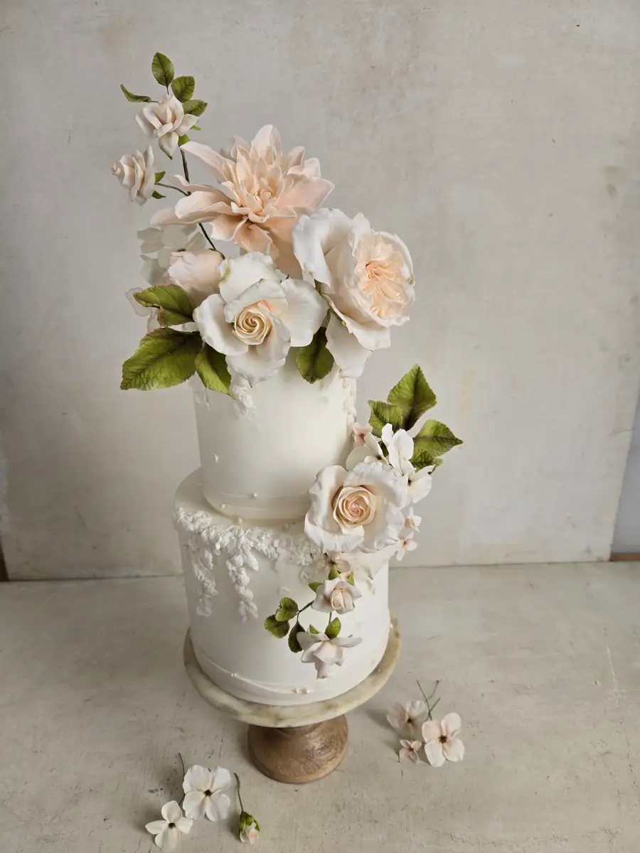 Two-tier white wedding cake with handmade peach and ivory sugar flowers on a wood stand
